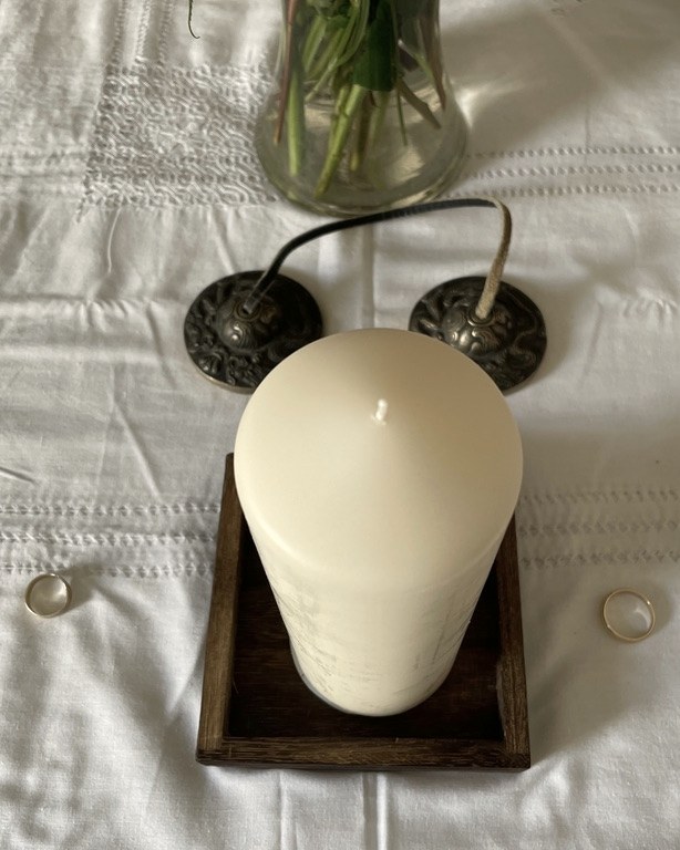 Two wedding rings on white linen, either side of a candle, with tingsha bells behind