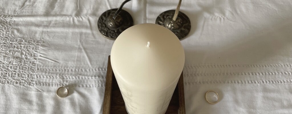 Two wedding rings on white linen, either side of a candle, with tingsha bells behind